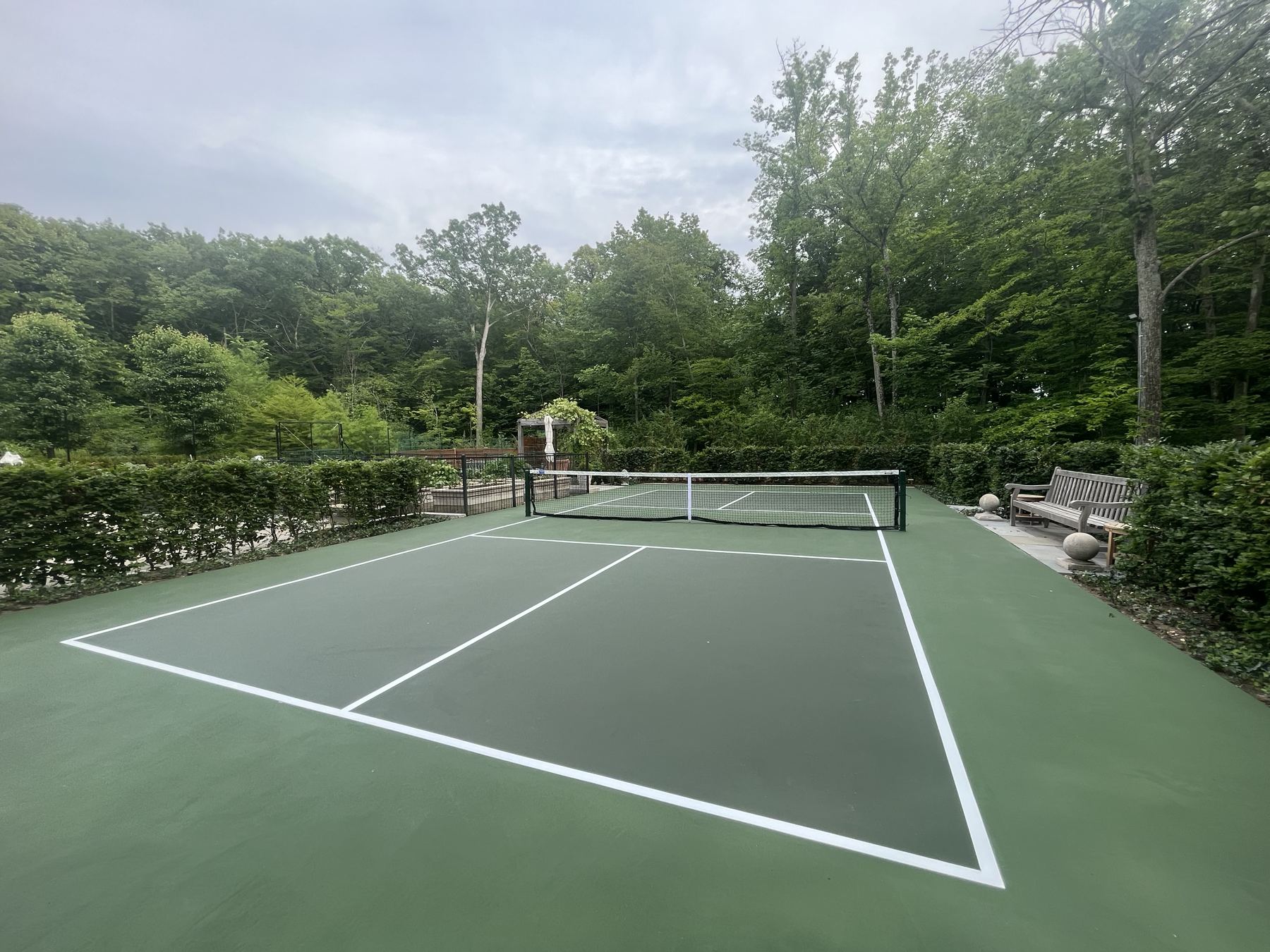 Dark green court framed by tall clipped hedges and mature woods, with a teak bench and stone patio